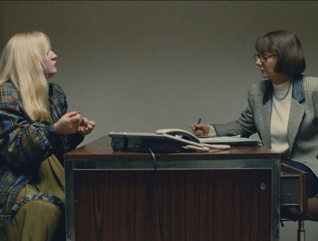 Two women, one in a suit, another in a dress and a jacket, sit across a desk from one another