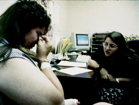 Grainy image of a white woman in tears as another white woman, in glasses, looks on from her desk