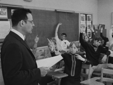 Black and white shot of a teacher addressing his students, whose hands are all up.
