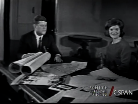 Black and white blurry C-SPAN screenshot of a man in a suit and a woman with fashionable hair seated at a table with paper plans in front of them