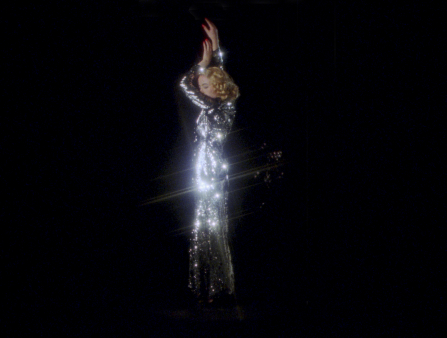 A white woman dances against a black background wearing a shimmery silver sequin dress