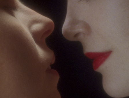 Tight close up of two white women one wearing bright red lipstick inching their faces closer together against a black backdrop