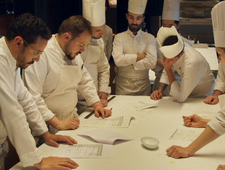 A group of chefs and kitchen staff nicely dressed in their whites gather around a table making notes ahead of service
