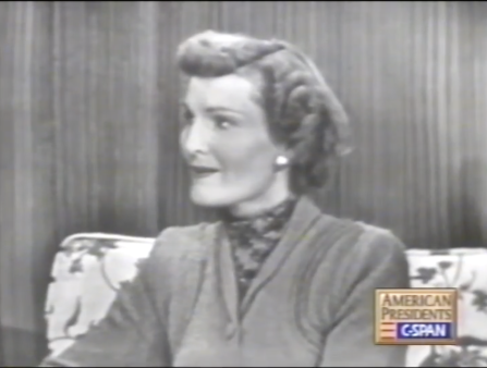 Black and white C-SPAN blurry screenshot of a white woman with tight curls and pearl earrings wearing a fashionable dress, seen from the waist up in a reading chair