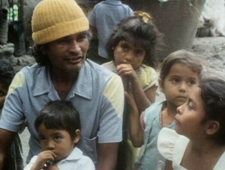 A brown-skinned man with a blue shirt and a yellow knitted-hat sits surrounded by young kids
