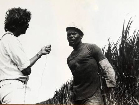Black and white image of a dark-skinned Black man in workwear and a construction hat being interviewed by a man dressed in all white with shaggy hair whose back is against the camera.
