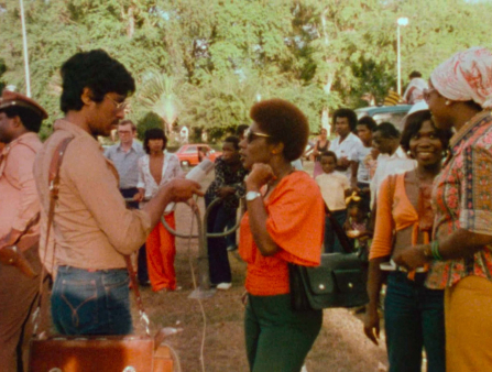 In an outdoor space surrounded by other people, a light-skinned man with dark short-cropped hair and gold glasses in a pink shirt holds a microphone up to a dark-skinned Black woman with a short afro and sunglasses in a bright orange shirt