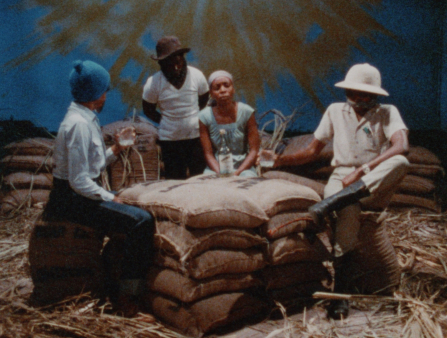 Four dark-skinned individuals (including a young woman in a blue dress and head wrap, and a man in hunting gear, including boots and hat) sit around a hay-filled floor drinking