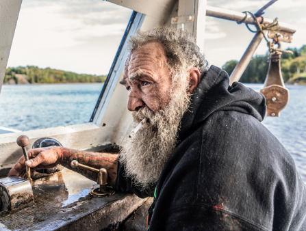 Gruff-looking older white man with frazzled grey hair and long haggard beard sits at his fishing ship, water in the background