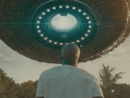 A balding Black man stares up at what looks like a UFO which is hovering right above him against a wild landscape