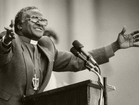 Black and white photo of an aging Black man wearing a priest's ensemble, arms open while speaking at a podium
