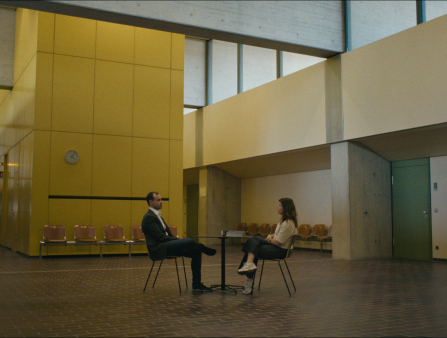 A middle-aged man and a woman sit across from one another in a yellow-walled open room