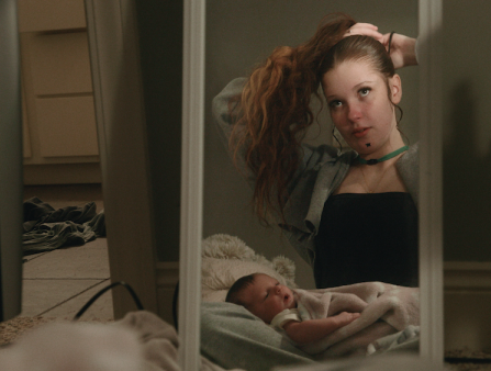 A young white woman with red hair is seen through a mirror tying her hair up as a baby lays on her lap.