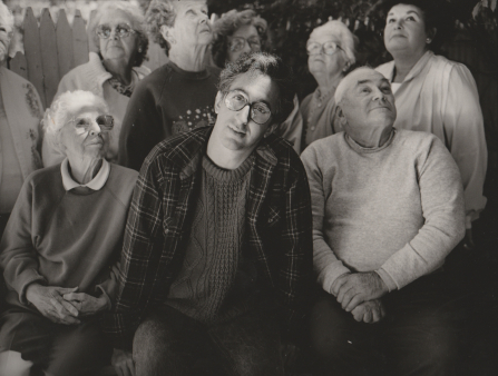 Black and white group shot of a middle-aged man in glasses, wearing flannel and a t-shirt surrounded by elderly men and woman all staring up above