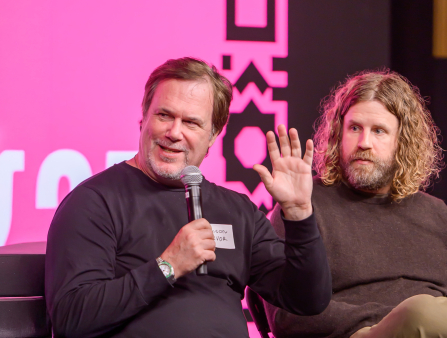 A middle-aged white man with short light brown hair, in jeans and a long-sleeved black shirt and boots sits on a stage holding a mic with a bright pink screen background behind him