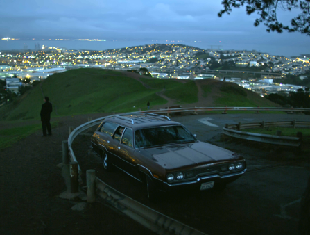 Scene at dusk; wide shot of a parked brown station wagon at an overlook, with a lit city down below, a lone figure standing still in the background.