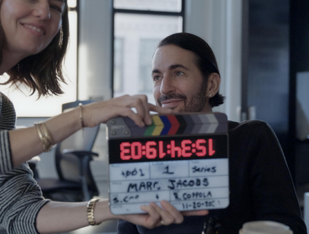 A middle-aged white man with slicked back black hair and five o'clock shadow sits in front of a camera as a white woman with shoulder-length brown hair holds a clacker that reads "Marc Jacobs" in front of him