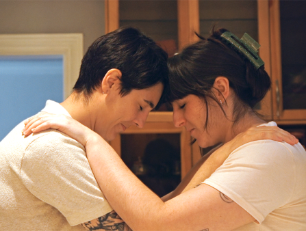 Two white women with dark hair (one cropped short, the other long with bangs, held up in a bun) wear white t-shirts as they embrace, their heads touching at their foreheads.