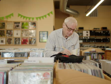 An elderly white man with thin white hair and glasses jots something on a notebook while in the middle of an old-school record store