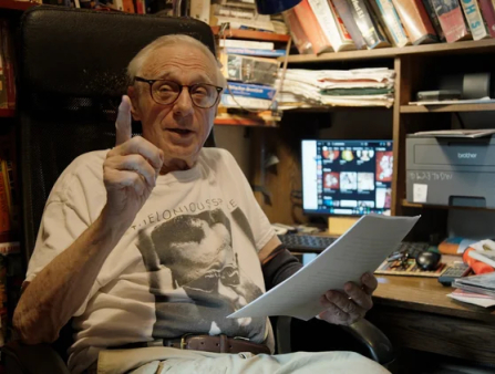An elderly white man with thinning white hair, dons glasses and a t-shirt while sitting at his crowded desk which brims with books and papers behind him