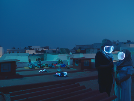 A blue-hued still of a couple in dystopian-looking hazmat helmets stadning atop a rooftop, as a group of individuals (sans masks) lay on roof in the background.