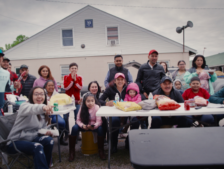 A large gathering of folks, both old and young, sits and stands looking at the camera in front of a table set up with food as a picnic.