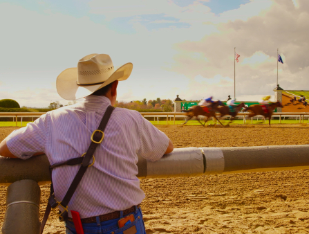 A man in a cowbow hat seen from behind is watching a horse race happening across from him
