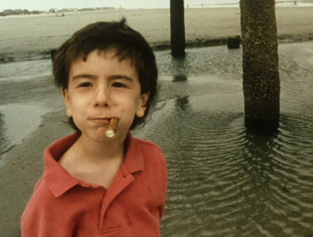 A young white boy with short black hair wears a red polo shirt while holding a pretzel in his mouth near a body of water