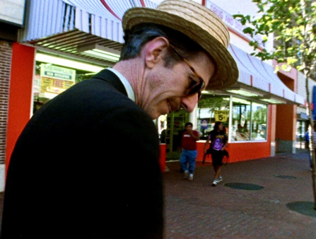 A tall white man wearing a straw hat, sunglasses and a blazer, looks down in profile out in the street.