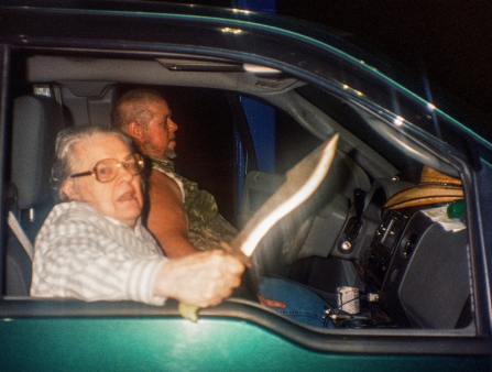 An older women with glasses sitting in a truck at night wields a knife at the camera