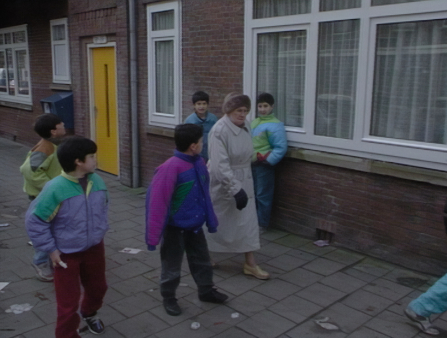An older white woman wearing a winter coat and hat passes a group of darker-skinned children on the street