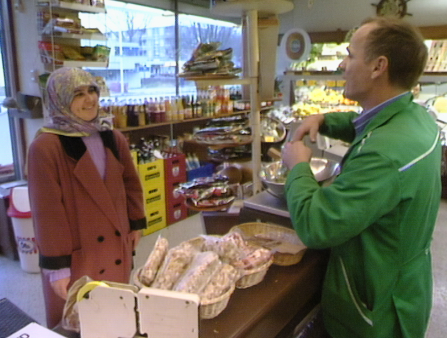 A woman wearing a hijab stands at the checkout counter at a small local shop, with a shopkeeper greeting her