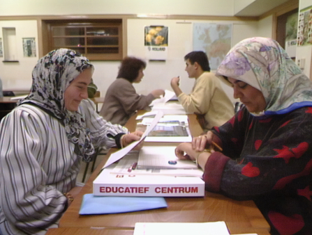 Two women wearing hijabs sit at a desk studying some papers in a public library
