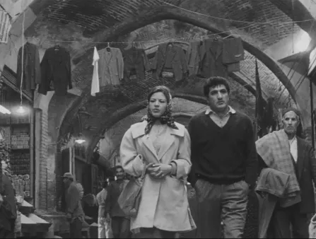 Black and white image of a woman in a coat and silk scarf over her hair walking alongside a man in pants ad a v-neck sweater around a Middle-Eastern market