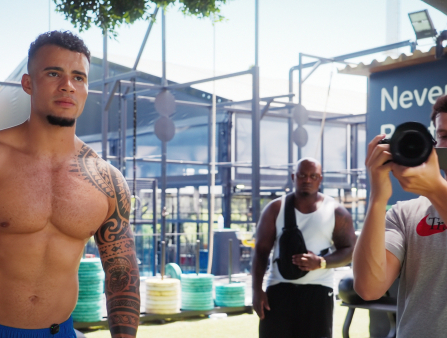A ripped, shirtless brown-skinned man with a shoulder/arm tattoo flexes as a young white guy in a t-shirt holds a camera aimed at us