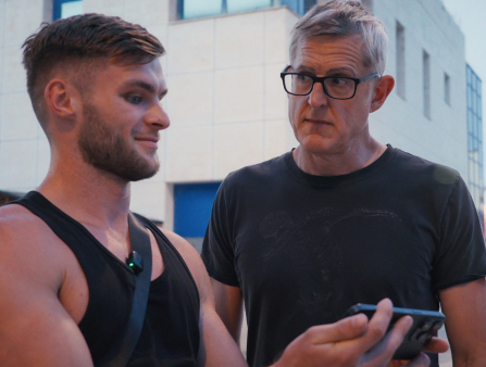 A middle-aged white man in a black t-shirt and rimmed glasses stares incredulously at a young muscled white man in a black tank top as he stares into his smartphone