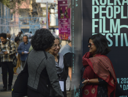 Three Indian women stand outside the street next to a banner promoting the Kolkata People's Film Festival