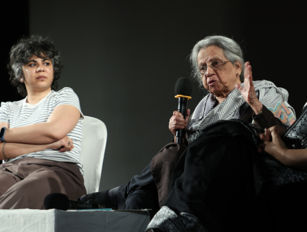 An aging Indian woman with grey haired pulled back sits at a stage, mic in hand, as another younger woman looks on.