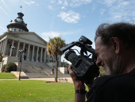 Wide shot of an older white man with long brown hair and beard holding a camera as he shoots a capitol building in the background of the shot against a sunny day