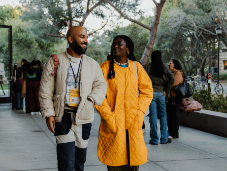 A bald Black male-presenting individual with a dark beard in a light jacket and funky pants walks outside next to a black woman with dark hair in a bright yellow jacket.