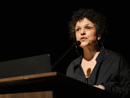 A middle-aged light-skinned Black woman with short curly hair wearing a black shirt stands at a lectern mid-speech