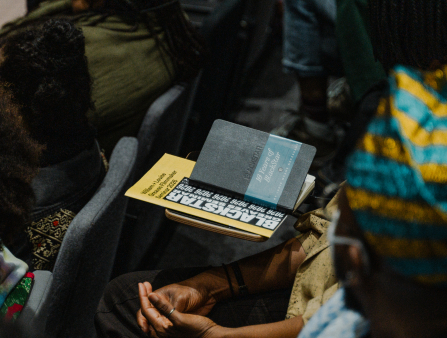 Close up shot of a Blackstar program and notebook on top of a desk, amid a number of Black folks seated as if for a lecture