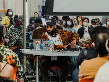 A Black male DJ in a Ford cap wearing an orange and black cardigan sits at a desk surrounded by folks in masks as they watch him mis something on his sticker-laden laptop