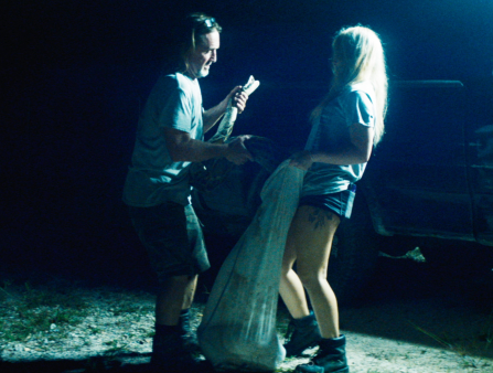 Nighttime shot of a middle-aged white man in jeans and a tee hoisting up a python into a bag held up by a young woman in a tee and shorts