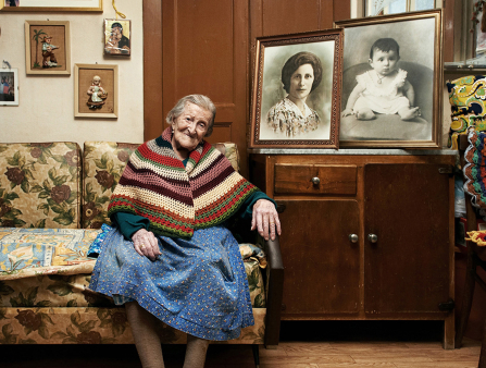 An elderly woman with pulled back grey hair sits on a yellow-print heavy coach surrounded by framed black and white photos of her younger self 