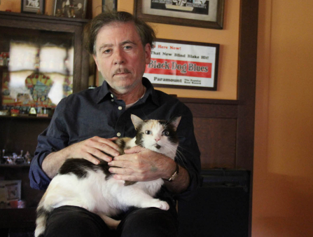 An older white man with thinning black hair and a bit of stubble, wears a dark blue shirt and dark pants as he sits staring at the camera as a white and black cat sits on his lap