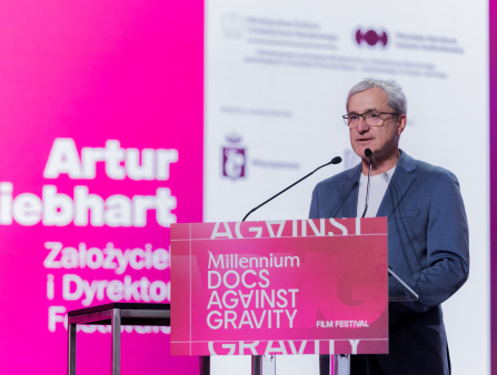 A middle-aged white man with grey hair and glasses wears a suit and a tee while standing at a podium that's branded as Millennium Docs Against Gravity in bright pink