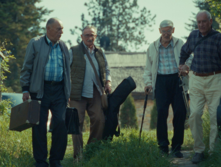 A group of elderly men in casual clothing carrying instruments in cases walk alongside a countryside road
