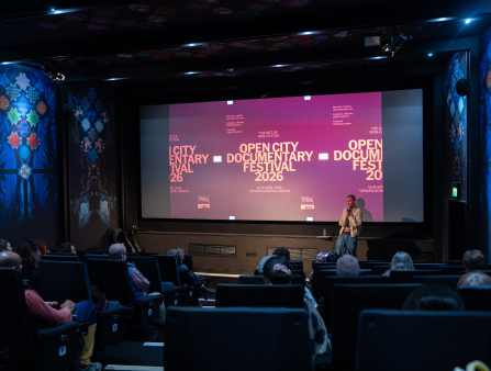 A white man stands in front of a crowded theater, a screen behind him showcasing an ad for the Open City Documentary Festival