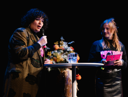A light-skinned woman with shoulder-length curly black hair in a stylish printed coat holds a mic at a table with two glasses as a young blond-haired woman wearing a CPH:DOX tee stands in the background